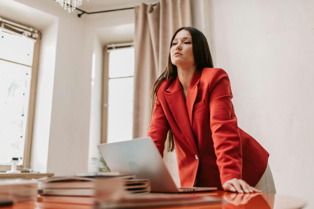 Businesswoman in a red blazer leaning over a desk with a laptop, exuding confidence.